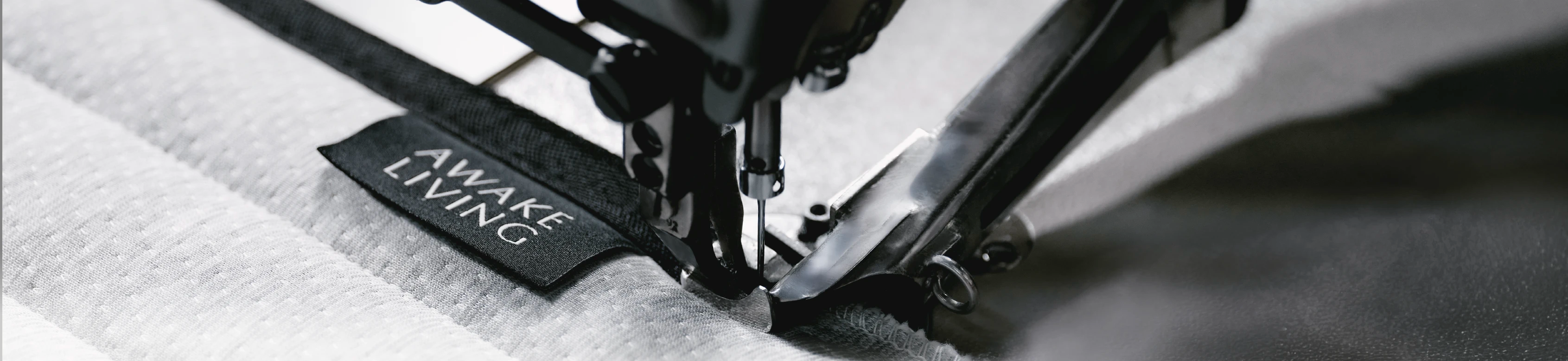 工業縫紉機的近距離特寫,展示機器正將印有 AWAKE LIVING 的黑色品牌標籤精準車縫至淺灰色的圍邊,展現細緻的製作工藝。
