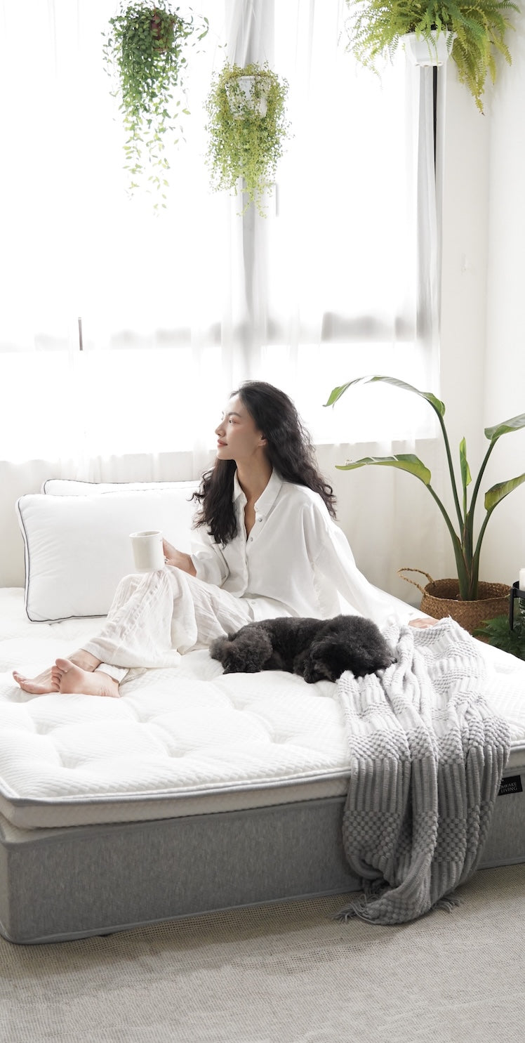 在充滿植物的明亮臥室裡,一位女性手持馬克杯坐在床上,一隻小狗睡在她腳邊。