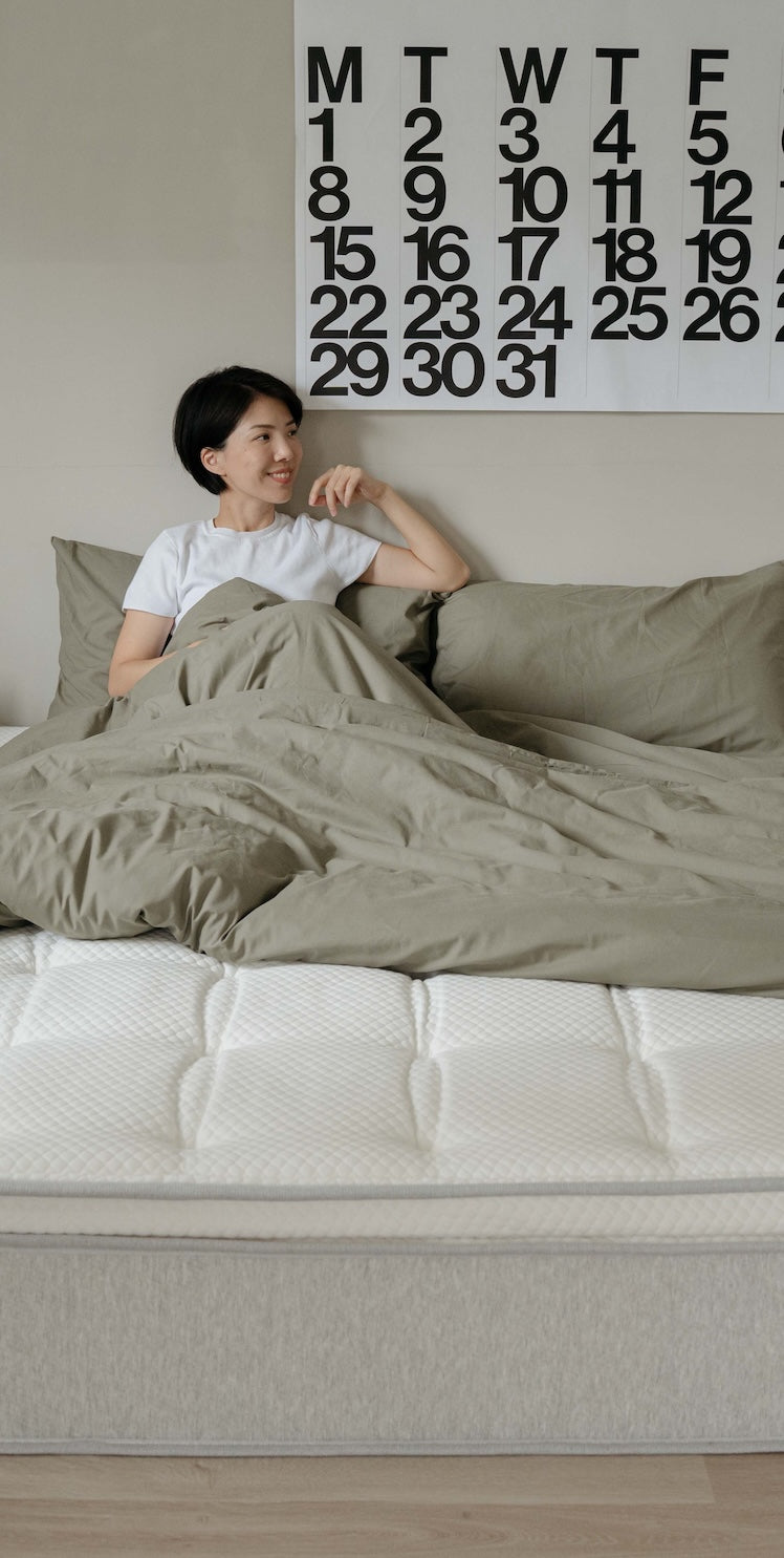 一位女性坐在床上,身後牆上掛著月曆,床尾露出白色床墊。