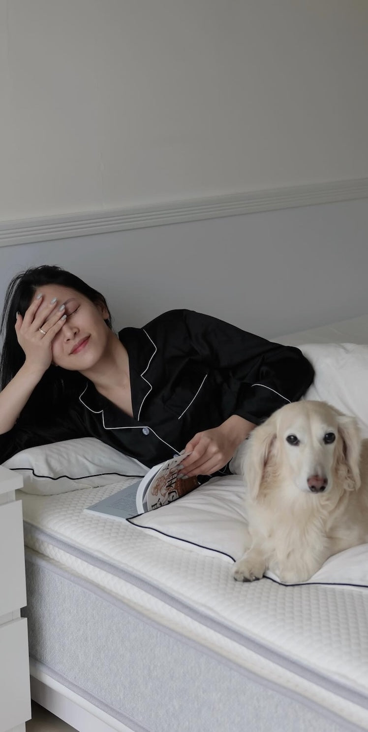 一位閉眼手撫額頭的女性,與一隻臘腸狗一同躺在床上休息。