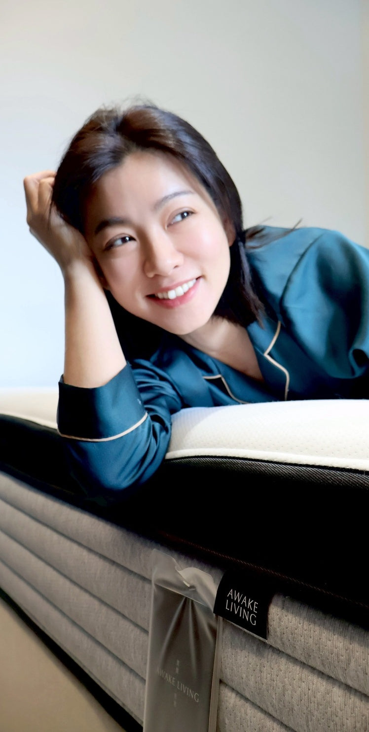 一位穿著緞面睡衣的女性,面帶微笑地趴在床墊上,前景是 Awake Living 品牌布標。