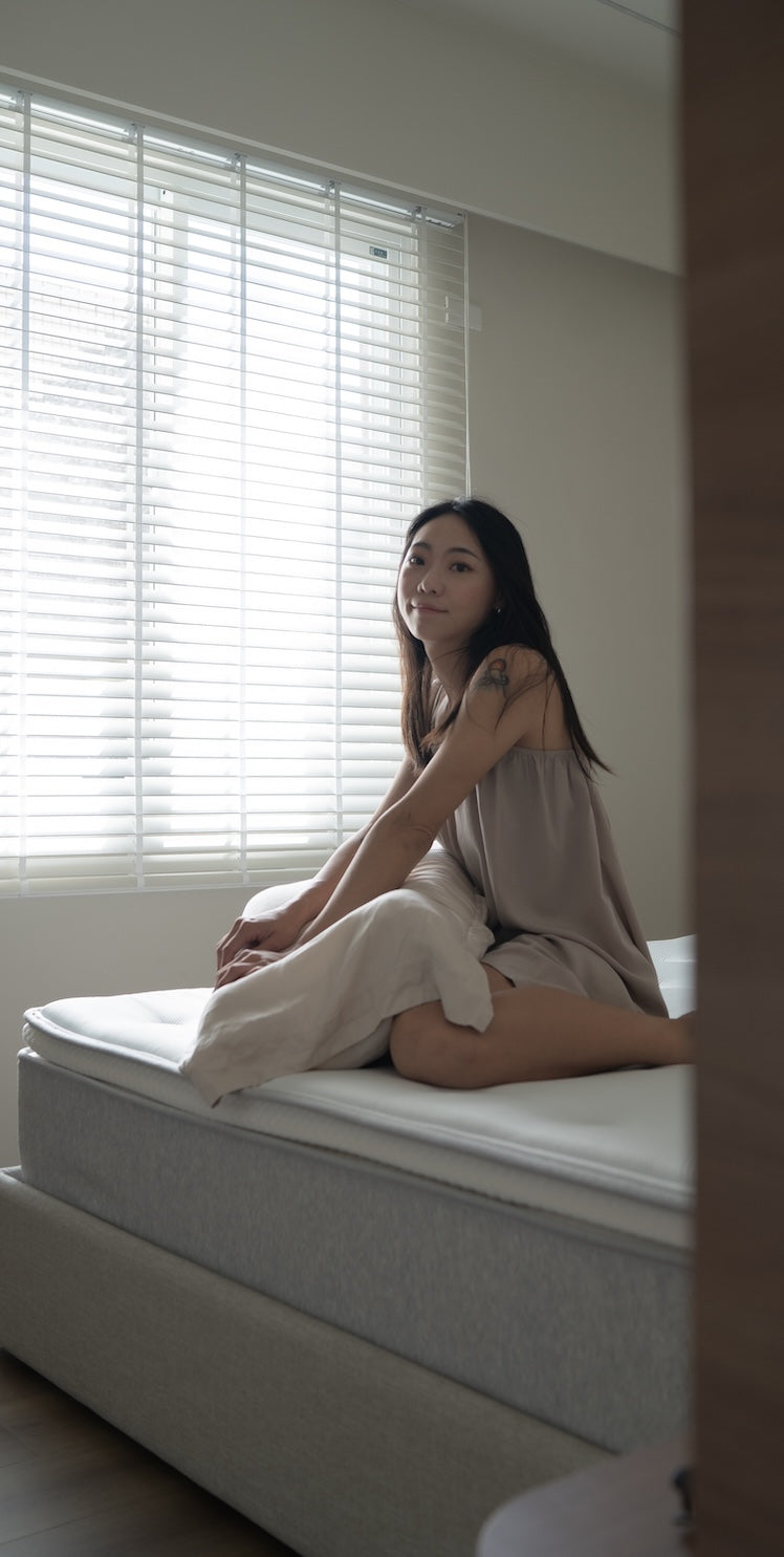 一位女性坐在床沿,身後是透著光的白色百葉窗,前景為門框。