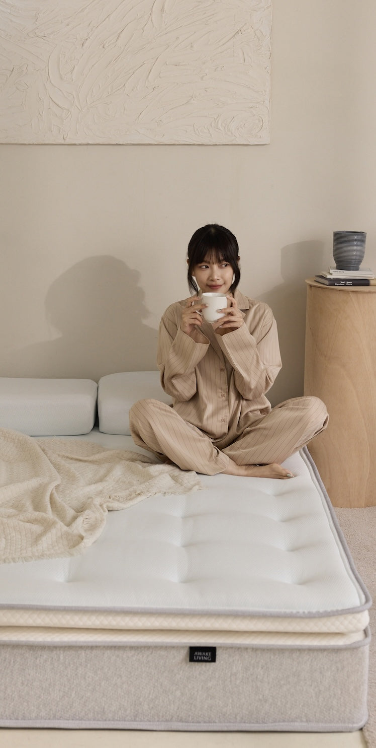 一位穿著睡衣的女性盤腿坐在床上,手持馬克杯,身後是兩個枕頭與牆面藝術品。
