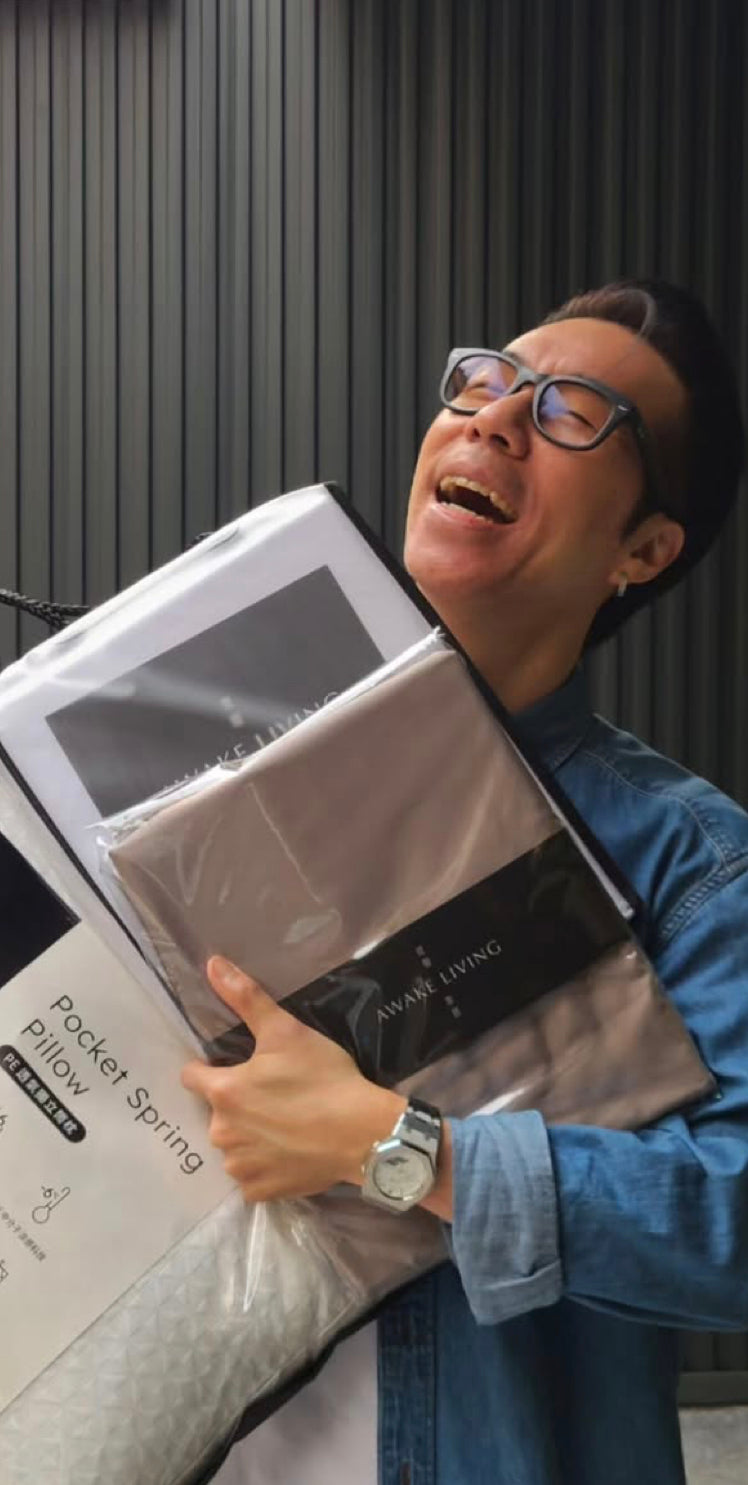 一位戴著眼鏡的男子開懷大笑,懷裡抱著一疊 Awake Living 的枕頭與寢具產品。