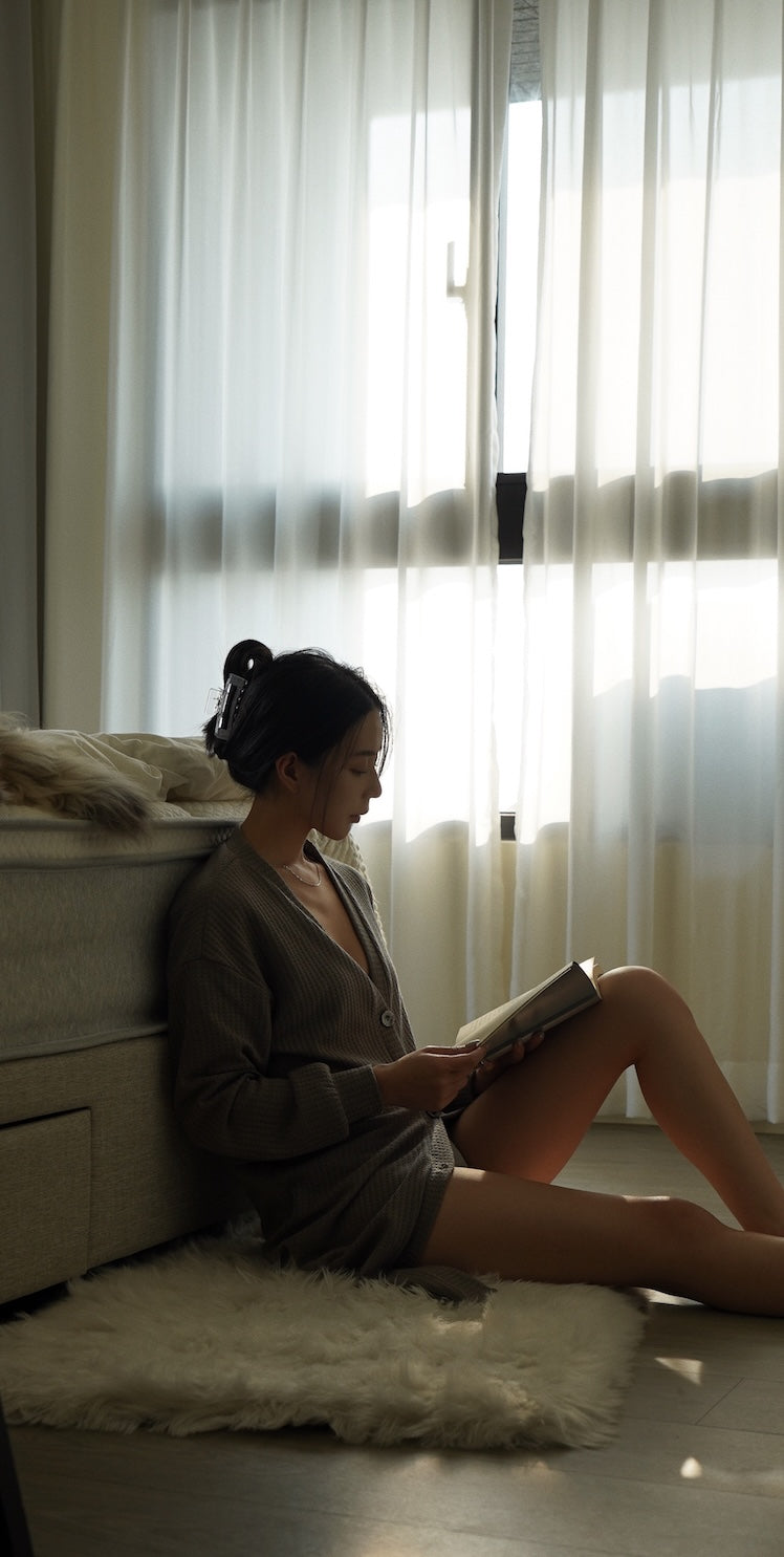 一位女性坐在地板的白色毛毯上,倚靠著床邊,在窗邊的光線下閱讀。