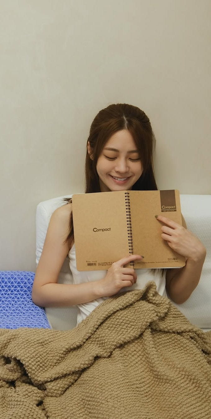 一位女性蓋著針織毯,面帶微笑地坐在床上翻閱筆記本。