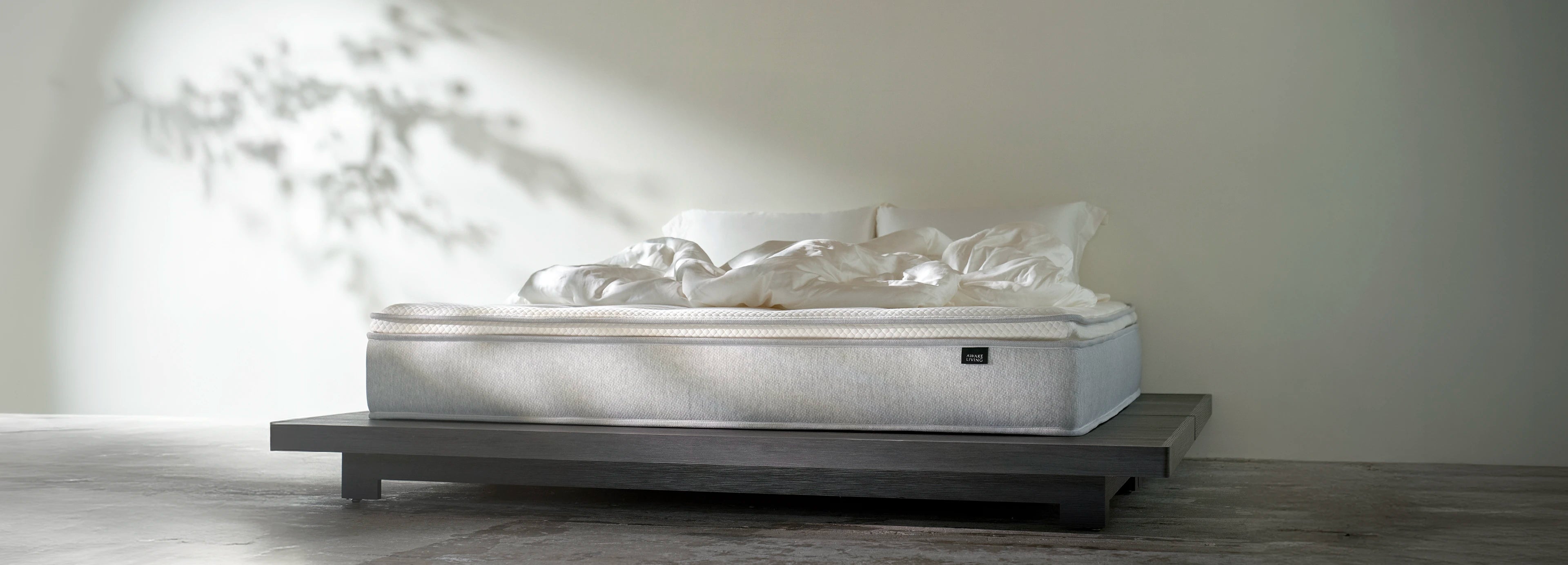 床墊放置在低矮的深色現代木質床架上，展示其使用情境。自然光線灑落，在背景牆上投射出清晰的樹影。