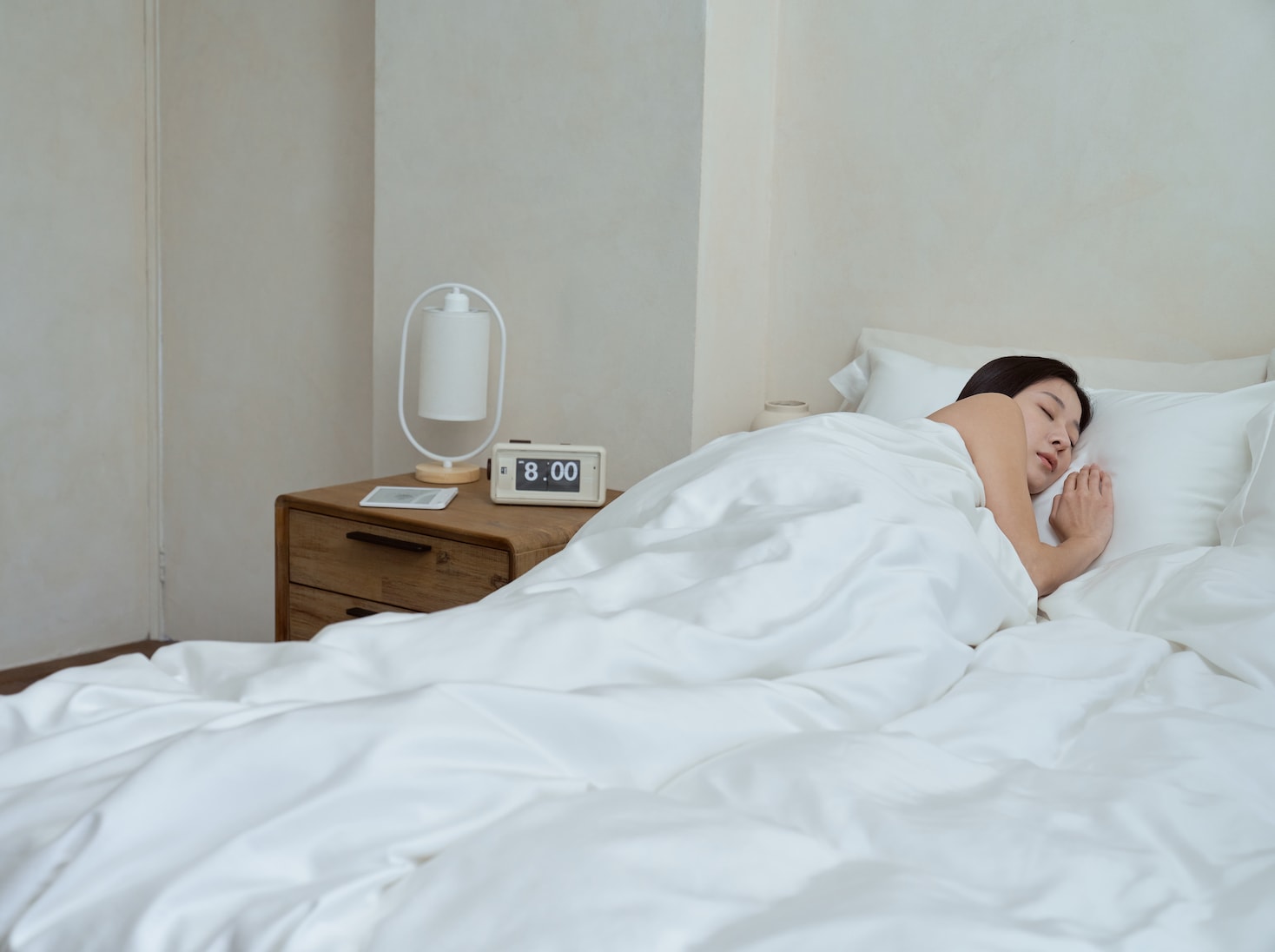 在一個明亮的早晨，一位女性睡在覺醒家居的 Original 經典好眠床墊上，床旁的茶几上放著鬧鐘和小檯燈，鬧鐘顯示著時間為 AM 8:00 - 桌機版本照片