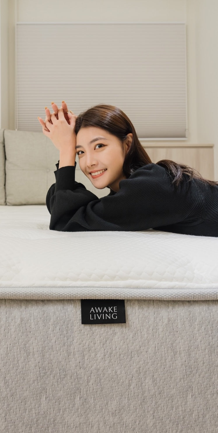 女子用手肘撐著自己身體俯在覺醒家居床墊上，帶著淺淺微笑看向著鏡頭方向