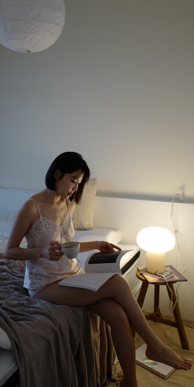 光線漸弱的房間，女子身著睡衣側坐在覺醒家居床邊，一邊翻著書本一邊喝著咖啡。